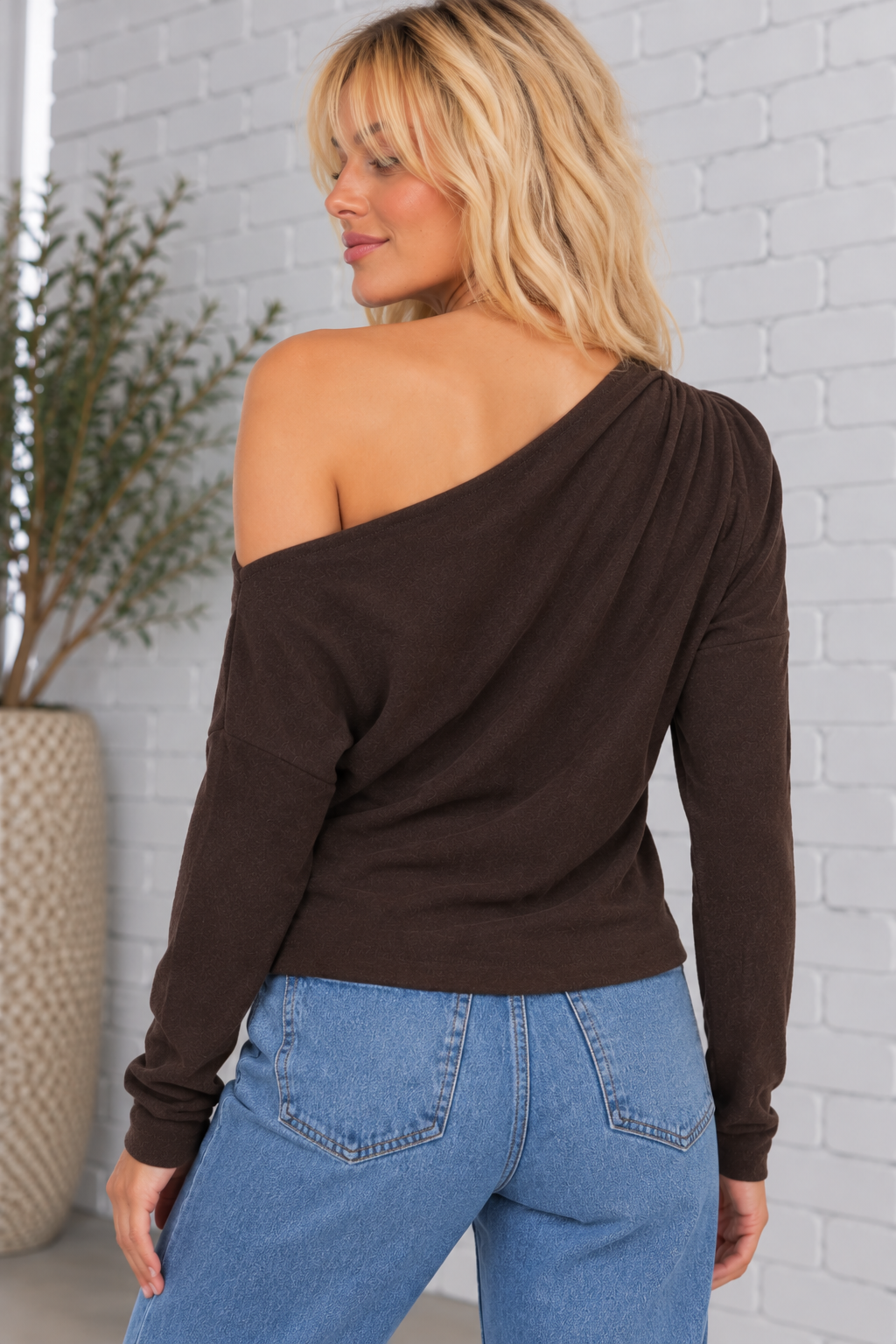 Back view of a model wearing the Colby Top in chocolate brown, featuring an asymmetrical neckline, soft draping, long sleeves, and a straight hemline.
