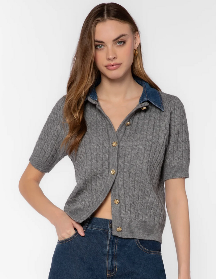 A woman modeling a short-sleeve gray cable-knit button-front cardigan with a blue denim collar and gold-tone buttons, paired with dark blue jeans against a plain light gray studio background.